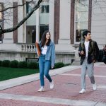Grupo de estudantes caminha por um passeio de um campus universitário num dia de sol.