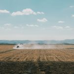 Trator levanta poeira enquanto trabalha num extenso campo agrícola sob céu limpo.