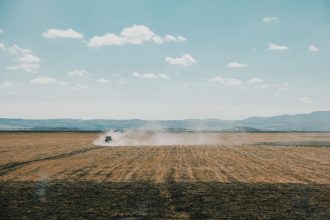 Trator levanta poeira enquanto trabalha num extenso campo agrícola sob céu limpo.