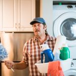 Homem idoso junto a uma máquina de lavar, segurando produtos de limpeza em casa.