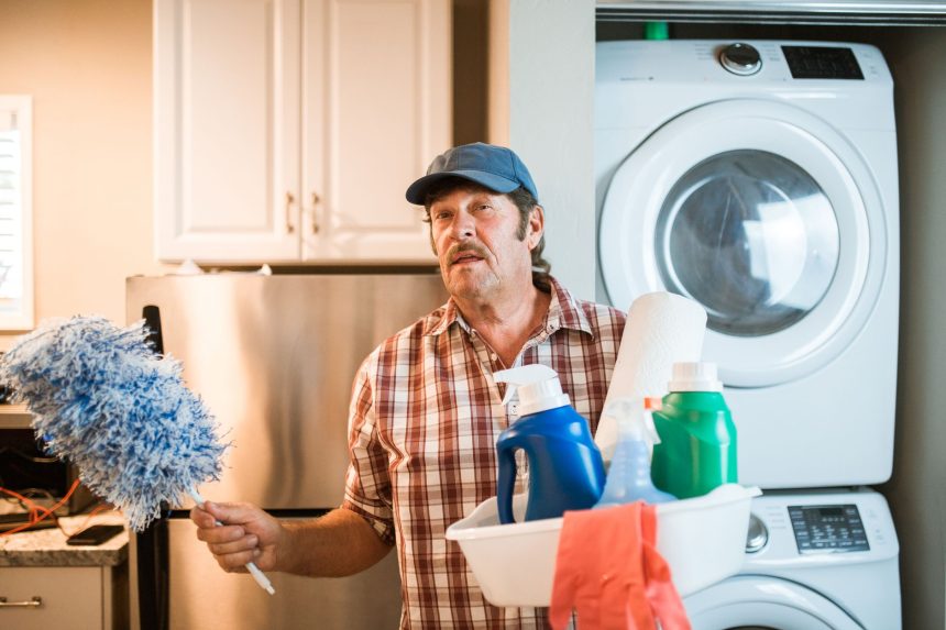 Homem idoso junto a uma máquina de lavar, segurando produtos de limpeza em casa.