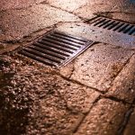 Grelhas de drenagem numa estrada urbana molhada, refletindo a luz durante a noite