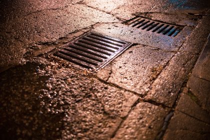 Grelhas de drenagem numa estrada urbana molhada, refletindo a luz durante a noite