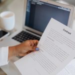 Pessoa segura um documento contratual junto a um computador portátil durante uma sessão de trabalho.