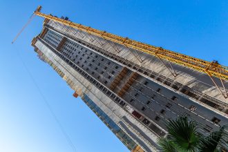 Gruas de construção sobre um edifício em obra, com a silhueta da cidade ao fundo, ilustrando o setor da habitação.
